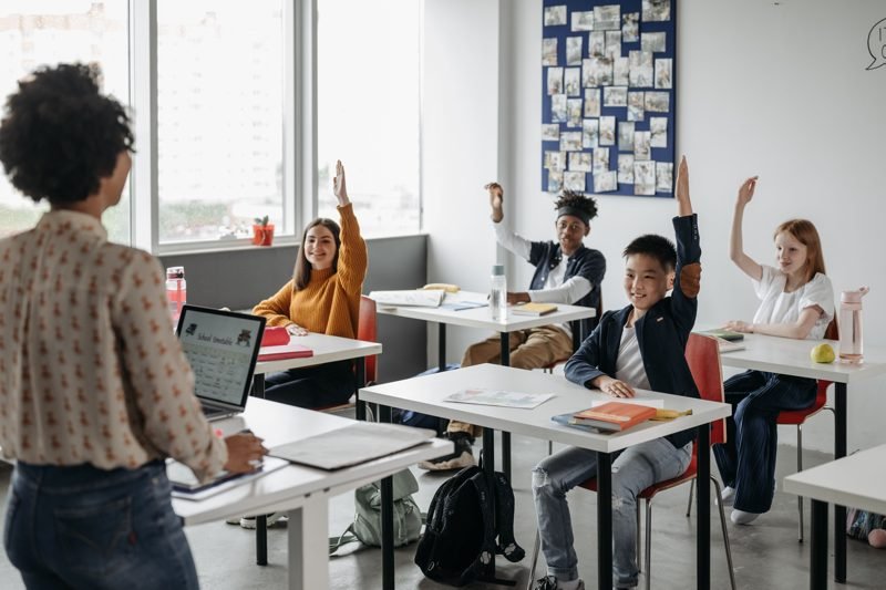 A teacher calls on students as four children raise their hands at their desks in a bright classroom.